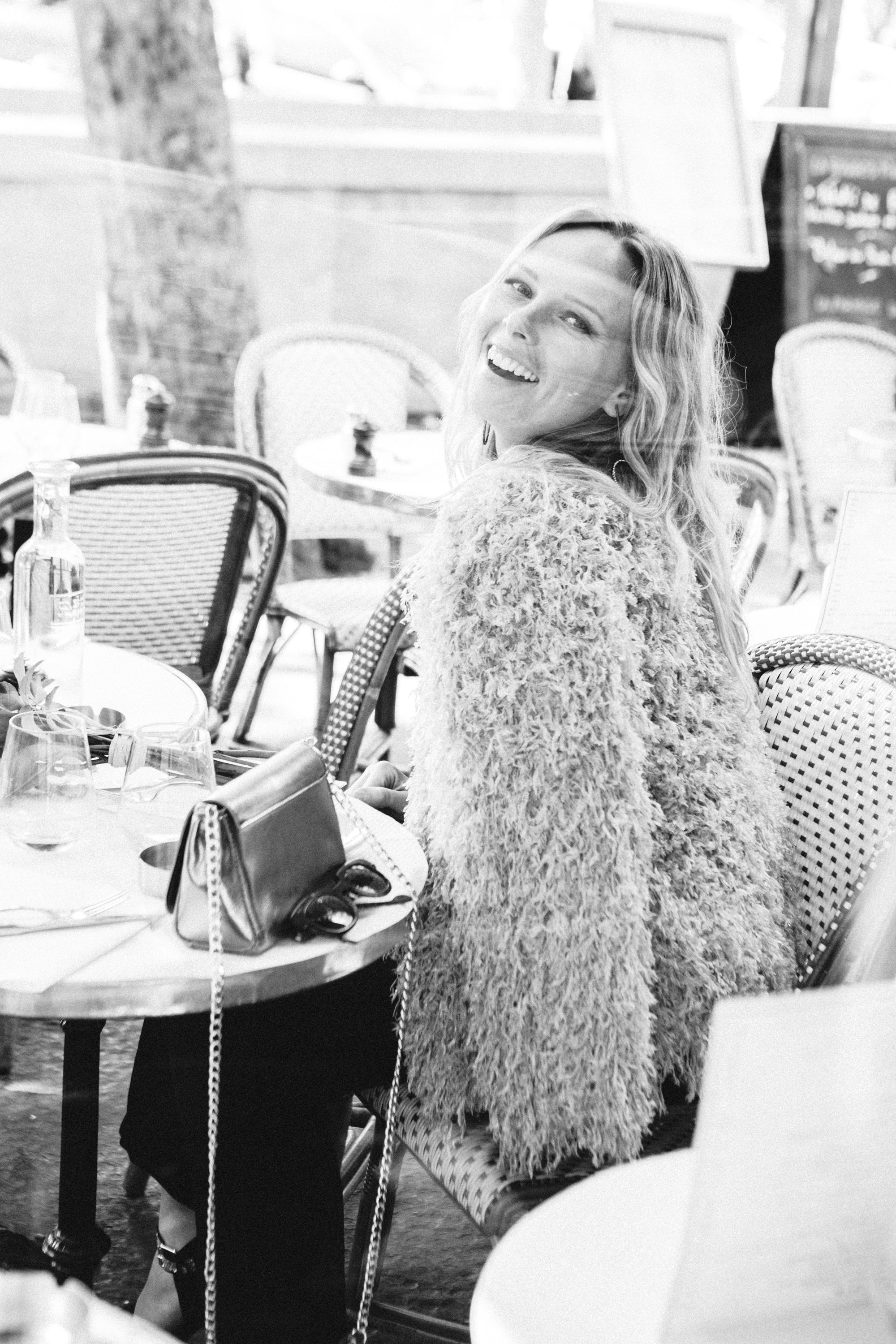 Woman sitting outdoors at a table with a blurred foreground, photographed through a cafe window, film noir style by Carla Coulson