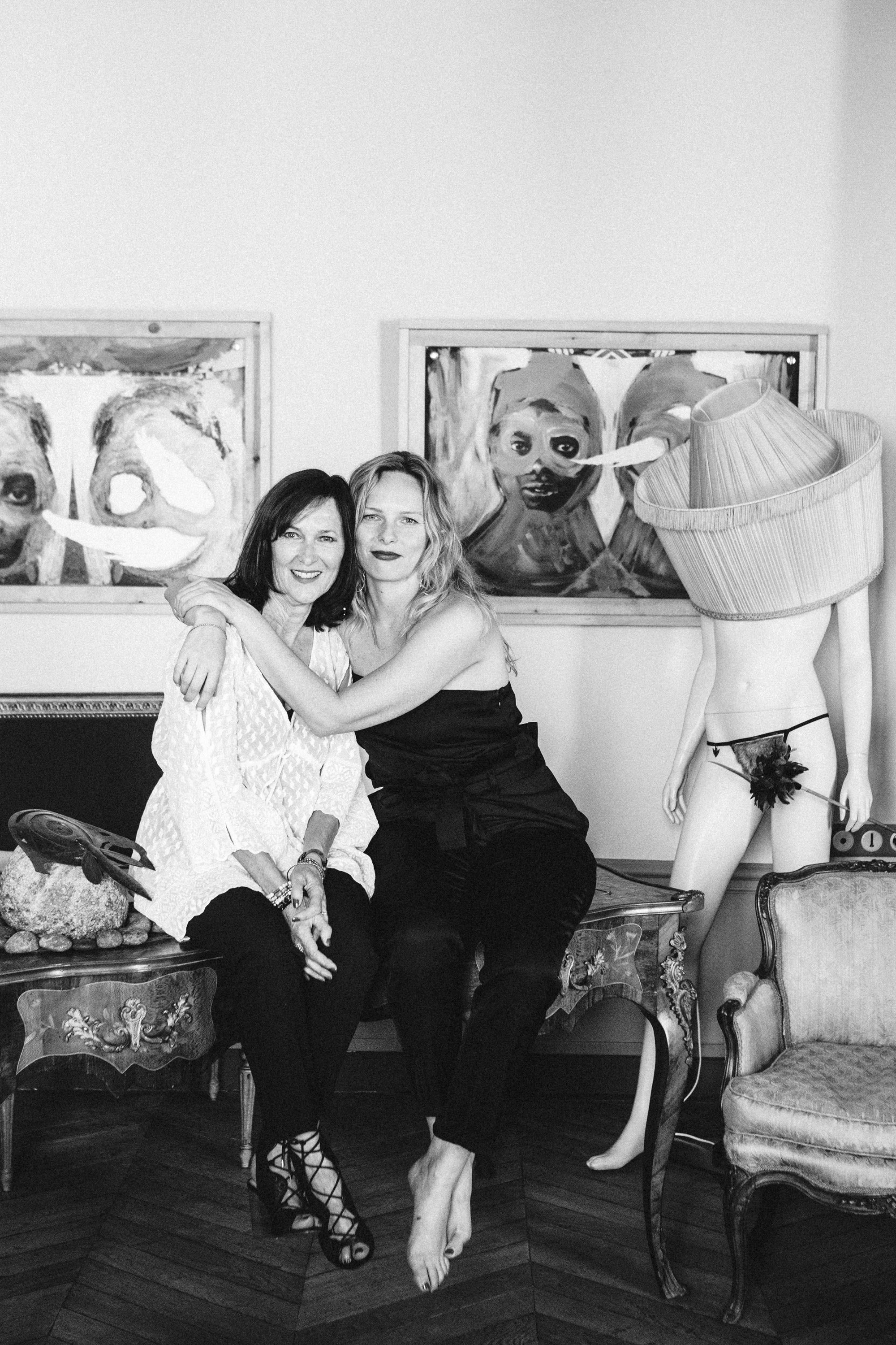Two women sitting on a couch with decorative items and mannequins in the background, Black and white film noir portrait - film noir photography by Carla Coulson