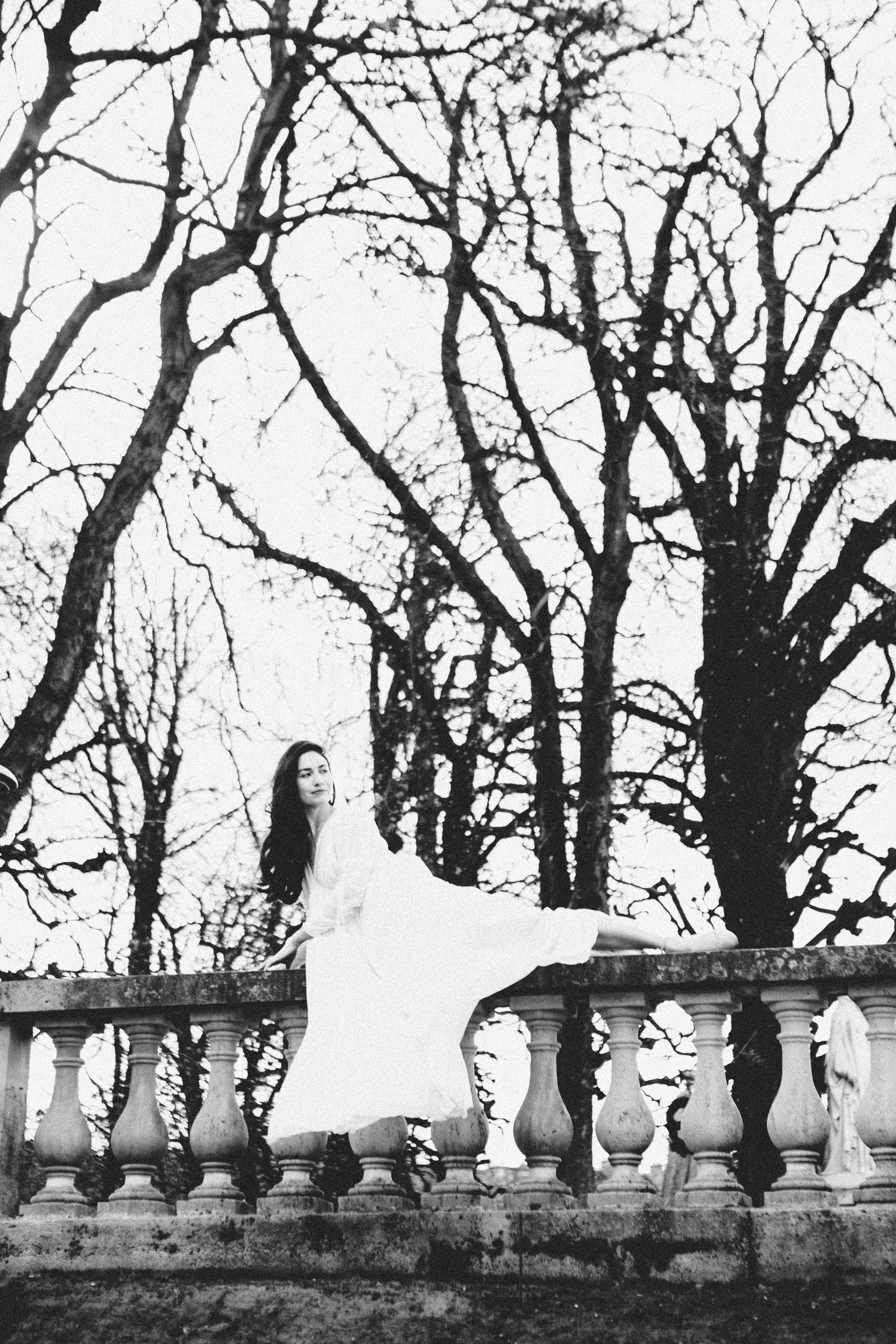 Woman in a white dress reclining on a stone railing with leafless trees in the background, film noir Paris portrait session by Carla Coulson