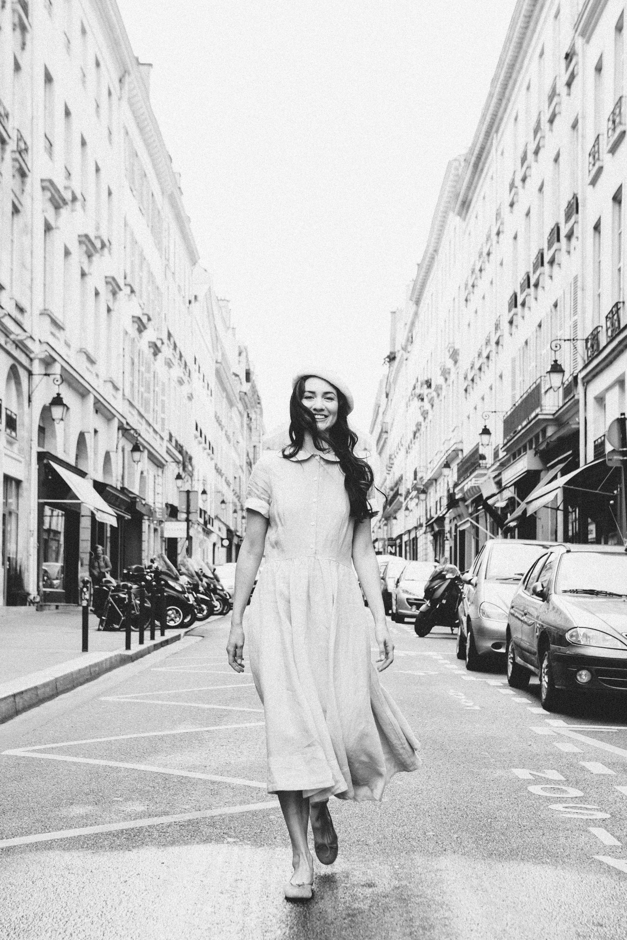 Young woman in a light dress walking down a Paris street with buildings and parked cars in the background, film noir portraiture by Carla Coulson