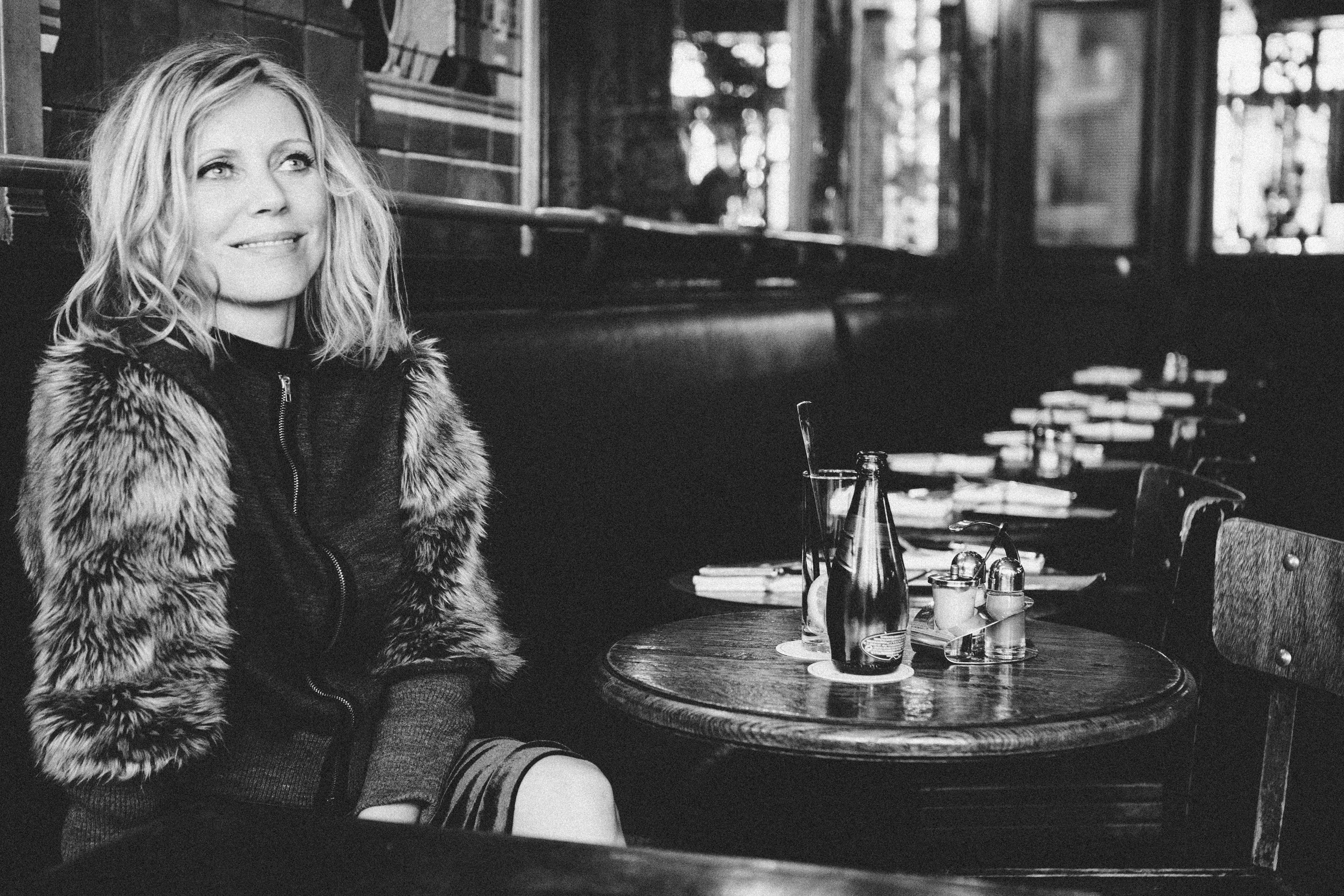 Woman sitting in a Paris cafe with a fur vest, surrounded by tables and chairs, dramatic black and white photography by Carla Coulson