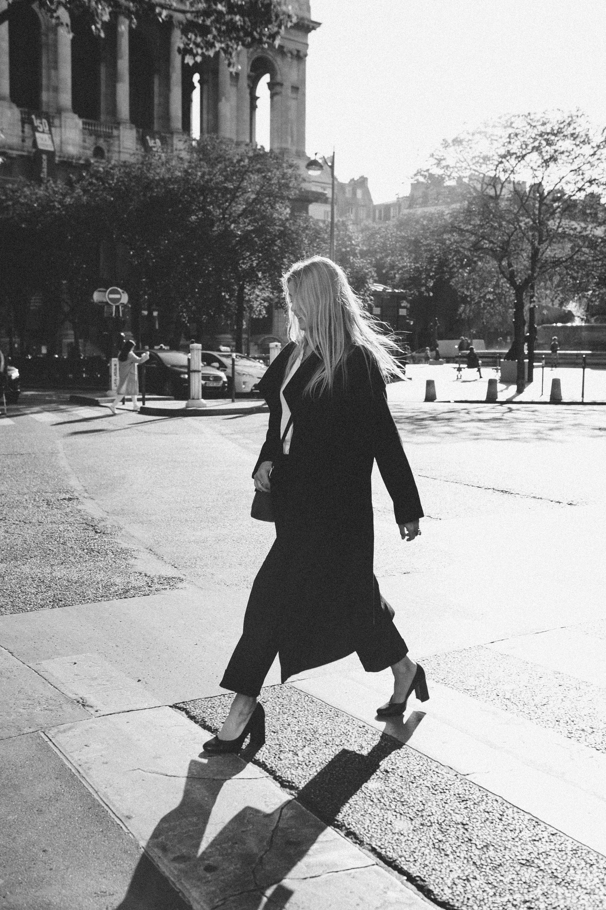 Woman in a black coat walking on a Paris street with trees and buildings in the background, intimate black and white photography by Carla Coulson