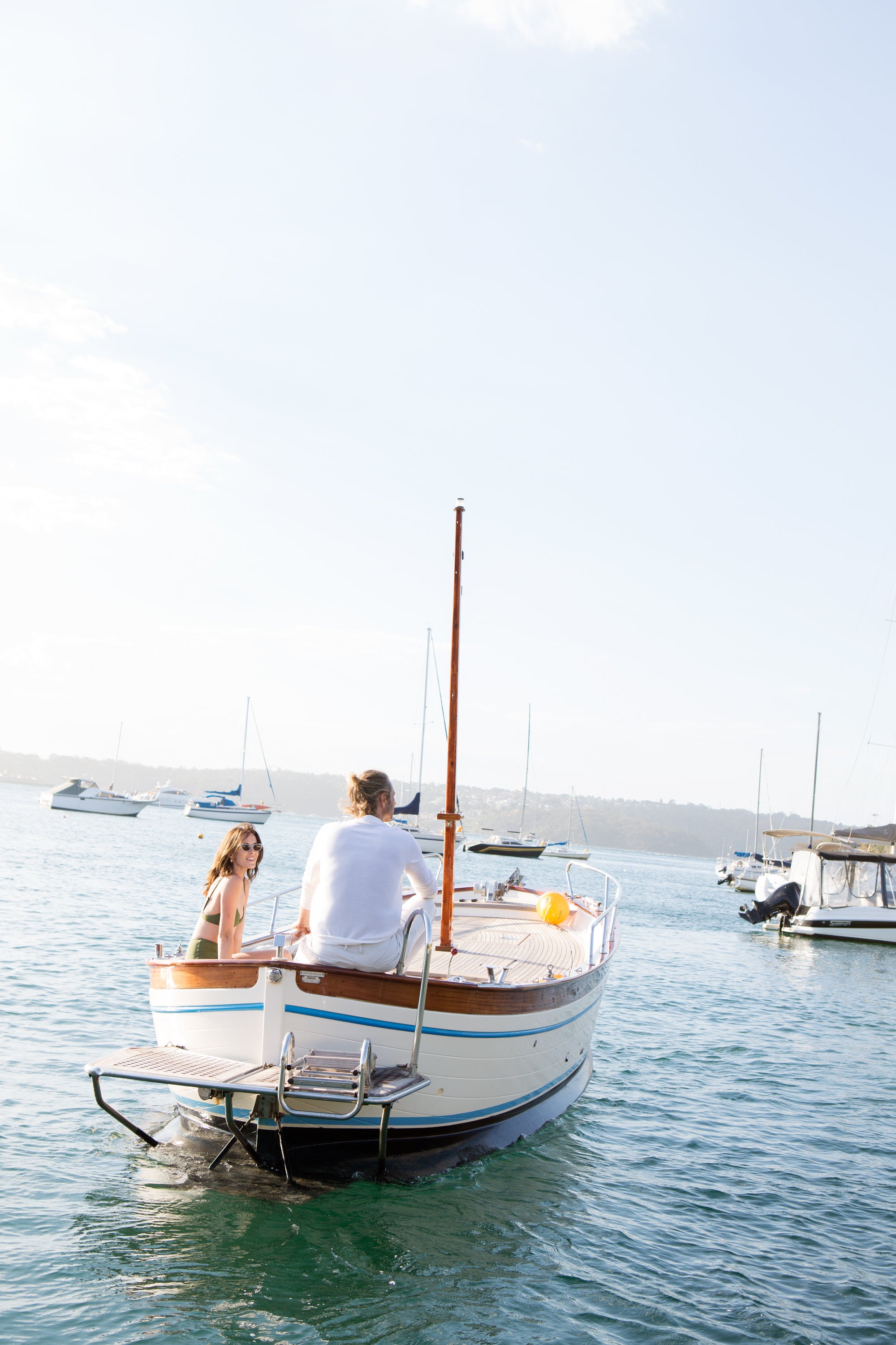 Two people on a sailboat in a harbor with other boats in the background — editorial photography by Carla Coulson